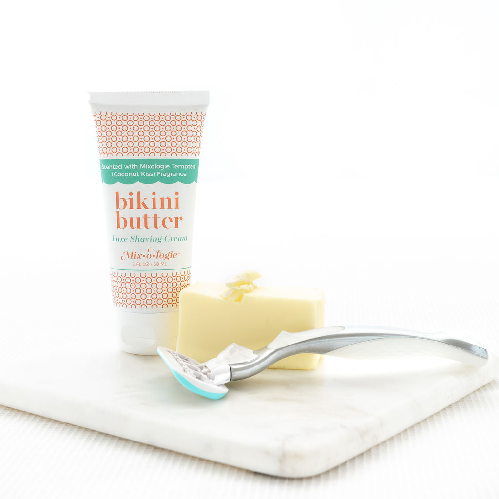 Luxe Shaving Cream Bikini Cutter scented with Mixologie Tempted (Coconut Kiss) Fragrance in a 2 fl oz or 60 mL white tube with orange circle pattern and white cap. Bikini Butter pictured with white background on marble board with razor and bar of soap.
